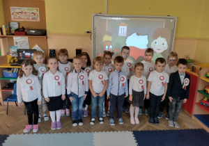 Grupa dzieci w strojach galowych stoi na tle symboli narodowych