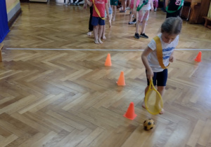 Dzieci za pomocą rakietki toczą piłkę slalomem do bramki